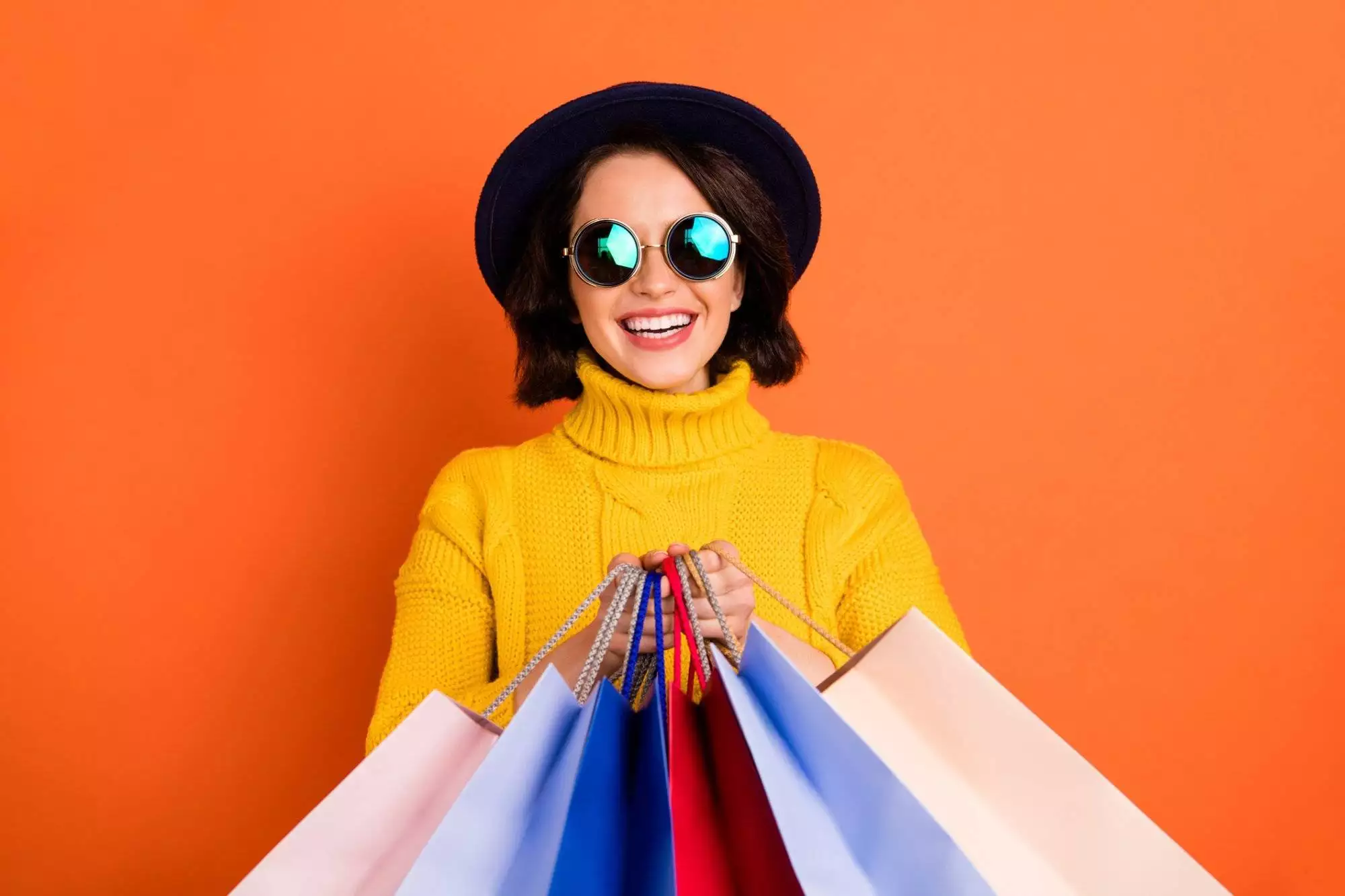 Photo of shopping cheerful girl wearing cap showing you what she has bought while isolated with orange background